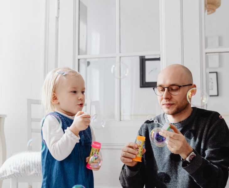 A joyful moment between a father and daughter blowing bubbles indoors, highlighting family fun.